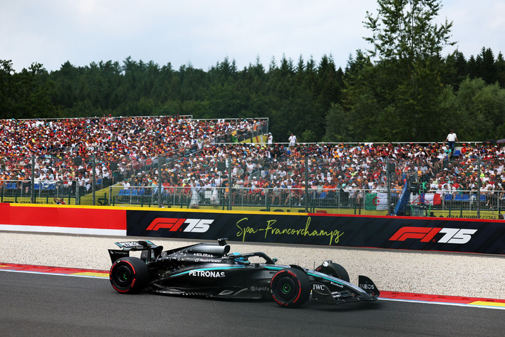 รถแข่งของทีม Mercedes-AMG PETRONAS Formula One Team กำลังวิ่งอยู่บนสนามสปา-ฟรังโกร์ชองส์ (Spa-Francorchamps) ด้านหลังมีอัฒจันทร์ที่เต็มไปด้วยแฟน ๆ ที่กำลังโบกธงจากหลายประเทศ โลโก้ Spa-Francorchamps ปรากฏอยู่บนแนวกั้น