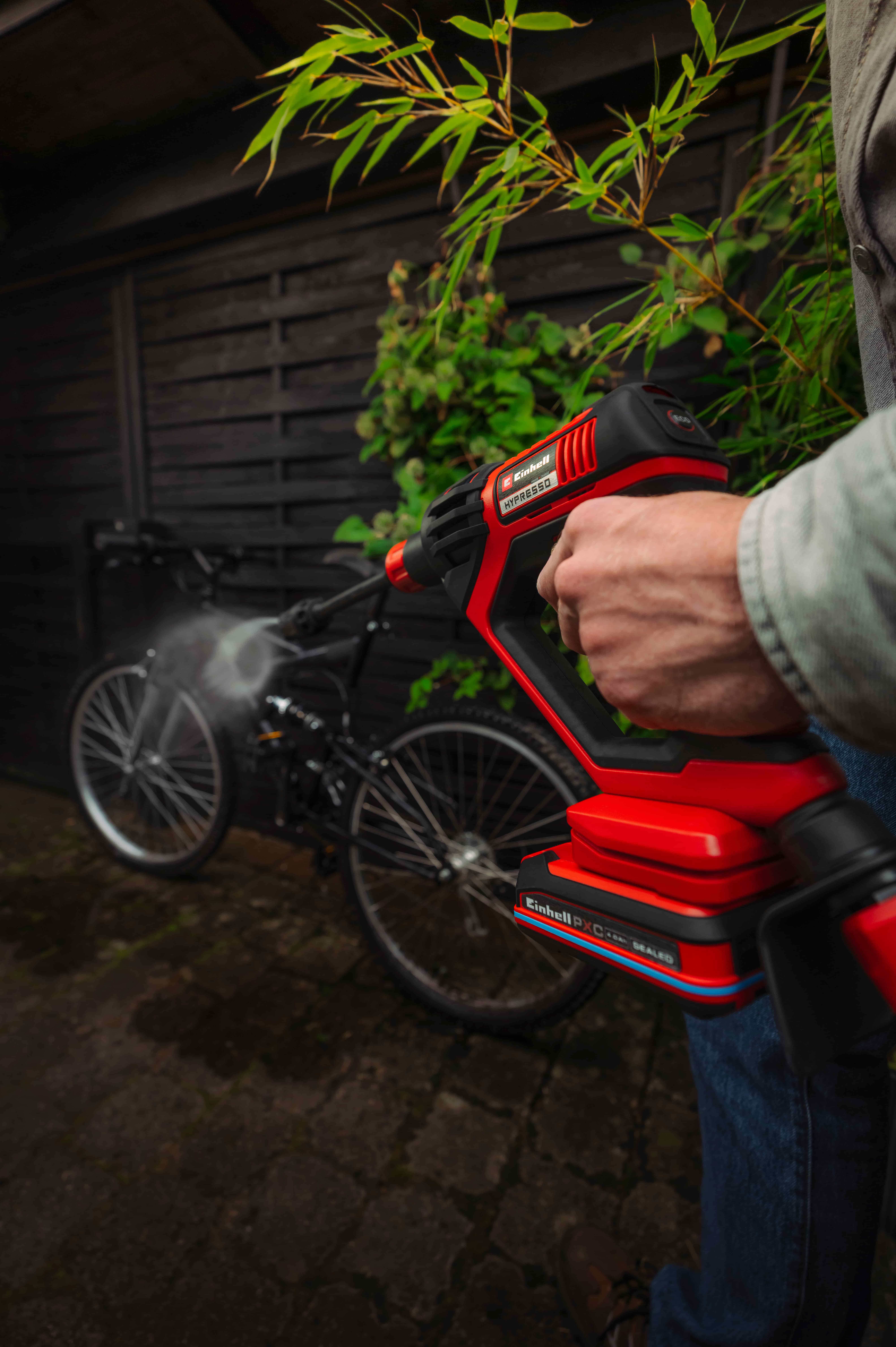 คนกำลังทำความสะอาดจักรยานด้วยเครื่องฉีดน้ำแรงดันสูงไร้สาย Einhell สีแดงในลานข้างกำแพงไม้