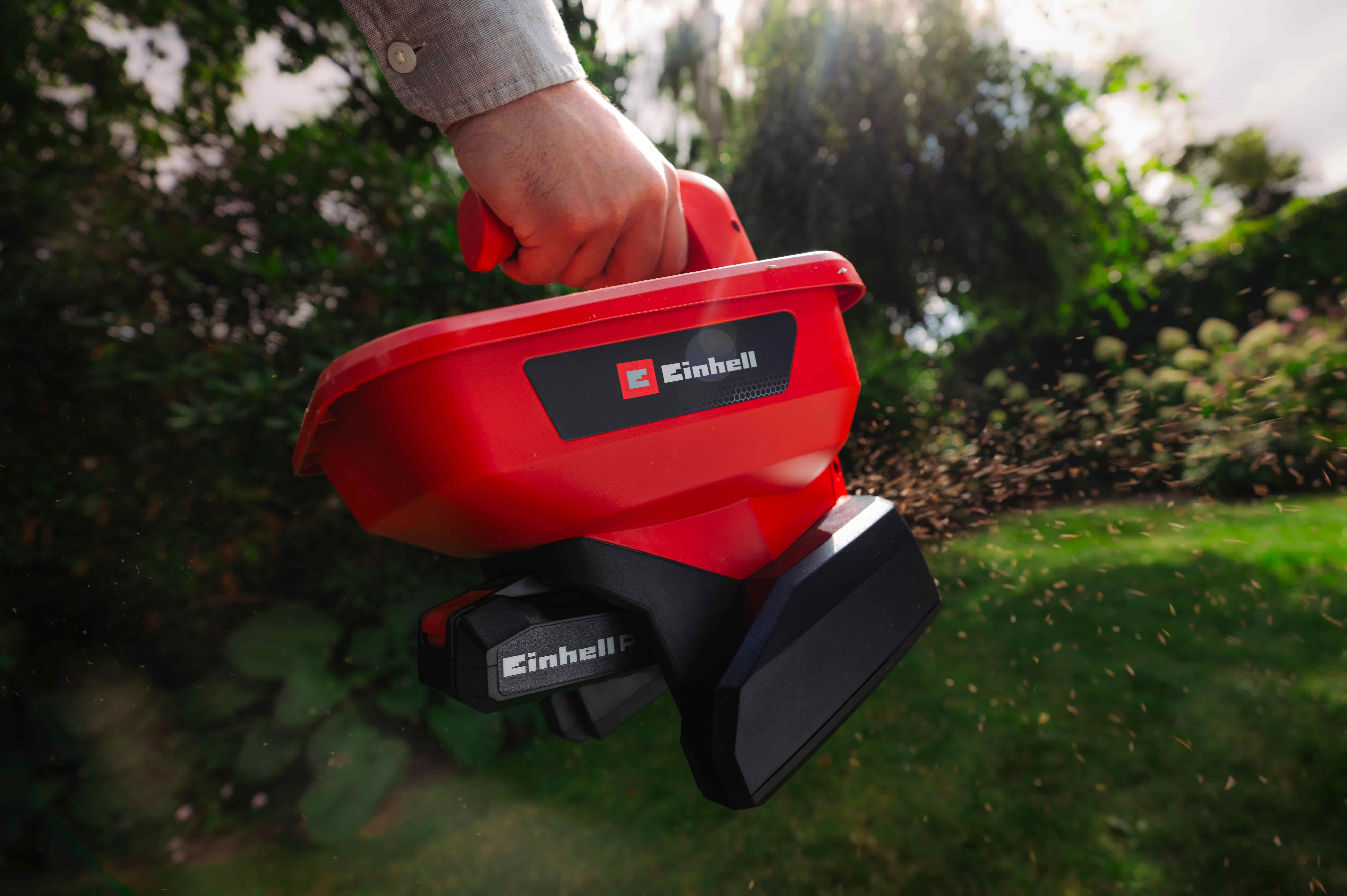 มือกำลังโปรยเมล็ดด้วยเครื่องหว่านเมล็ดไร้สาย Einhell สีแดง ขณะที่เมล็ดเล็ก ๆ กระจายไปทั่วสวน