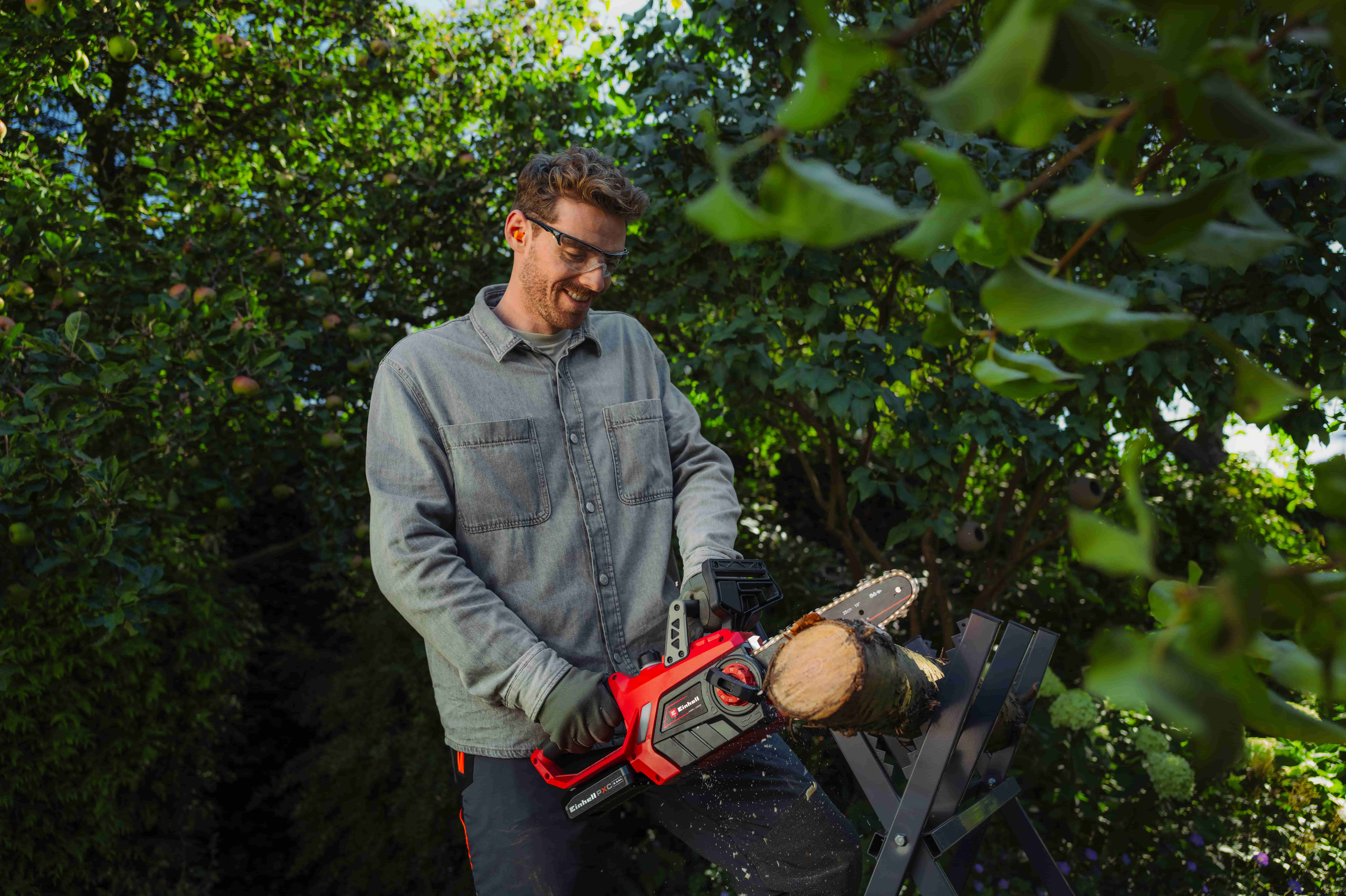 ผู้ชายสวมแว่นตานิรภัยกำลังตัดท่อนไม้ด้วยเลื่อยโซ่ยนต์ไร้สาย Einhell สีแดง บนแท่นเลื่อยในสวน