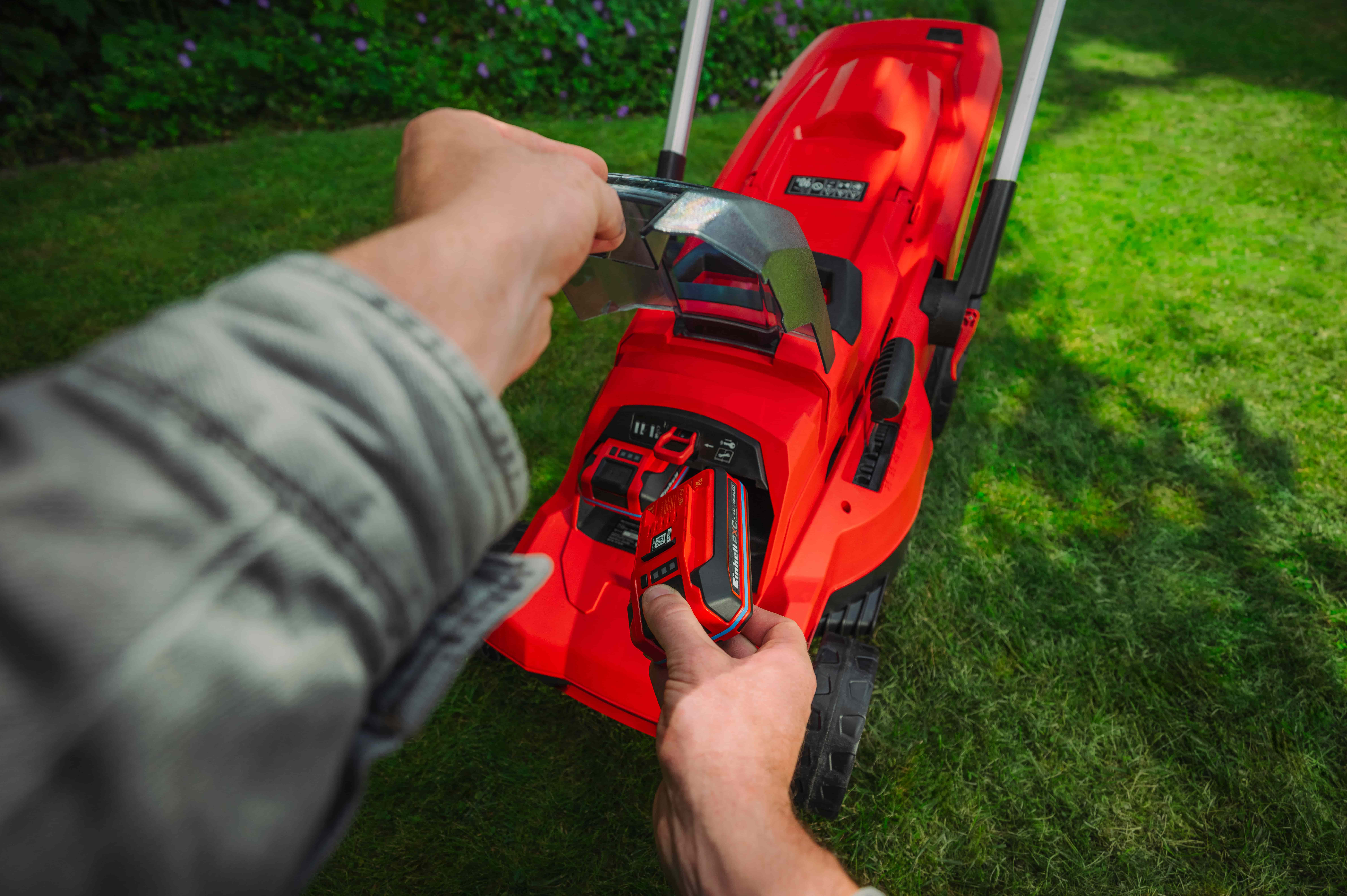 มือกำลังใส่แบตเตอรี่ Einhell Power X-Change เข้าไปในเครื่องตัดหญ้าไร้สาย Einhell สีแดงบนสนามหญ้า