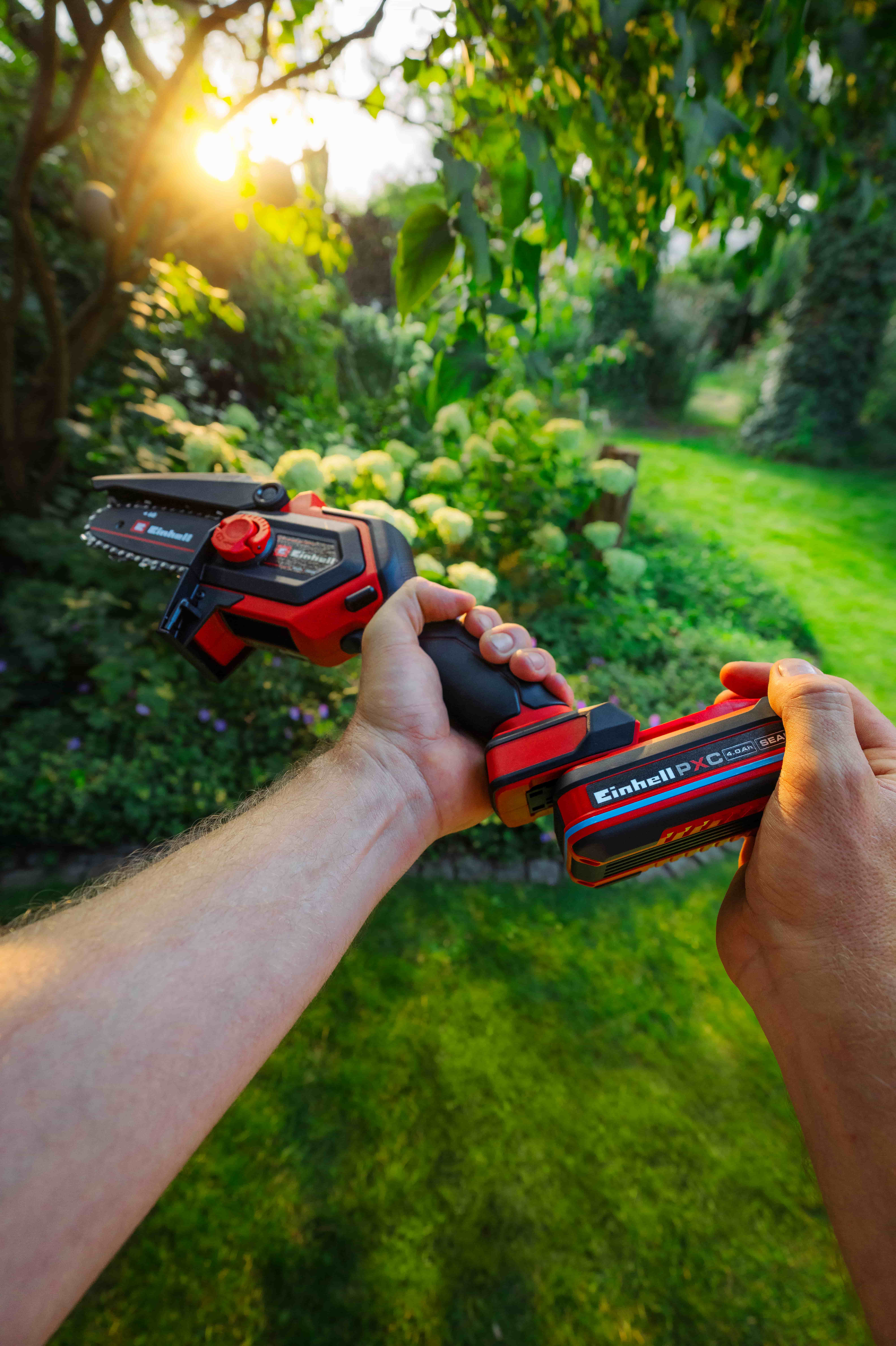 มือกำลังใส่แบตเตอรี่ Einhell Power X-Change เข้าไปในเลื่อยโซ่ยนต์ไร้สาย Einhell สีแดงขนาดเล็กในสวน